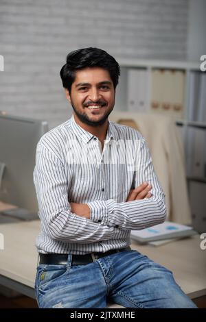 Confident young Indian businessman sitting on edge of his table and looking at camera Stock Photo