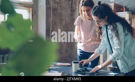 Pretty young women are standing near table, placing colorful pictures on it and shooting flat lay with smartphone. They are sharing ideas about design and smiling. Creative teamwork concept. Stock Photo