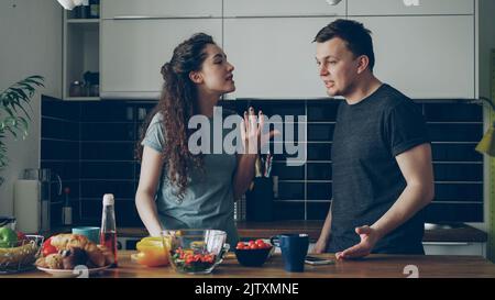 Pretty woman standing at table cutting salad while her boyfriend comes and they argueing speaking loud and nervous. Relations , difficulties concept Stock Photo