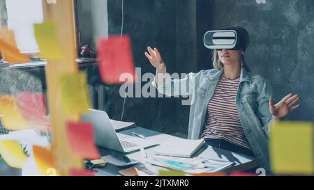 Attractive young woman is sitting at table and wearing virtual reality glasses. She is moving hands and and having VR experience in modern lof office indoors Stock Photo