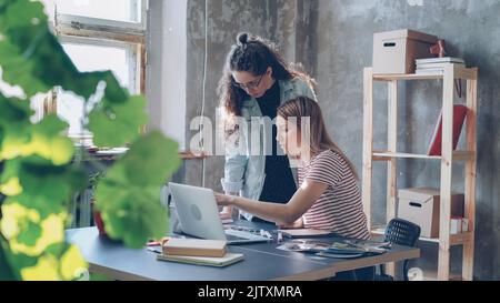 Two attractive colleagues are discussing design watching notes in notebook, laptop screen and photos on table. Women are talking and gesturing emotionally, they are involved in project. Stock Photo