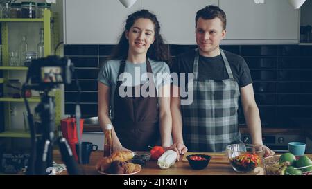 Smiling attractive couple recording video food vlog about cooking haelthy salad on digital camera in the kitchen at home. Vlogging and social media concept Stock Photo