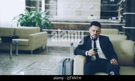 Pan shot of concentrated businessman using smartphone sitting on armchair luxury hotel with luggage near him Stock Photo