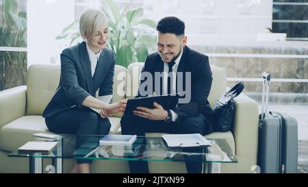 Two female and male business colleagues using digital tablet, notepad and talking at hotel lobby while sitting on couch and waiting check in at reception Stock Photo