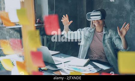 Attractive young woman is sitting at table and wearing virtual reality glasses. She is moving hands and and having VR experience in modern lof office indoors Stock Photo