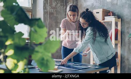 Pretty young women are standing near table, placing colorful pictures on it and shooting flat lay with smartphone. They are sharing ideas about design and smiling. Creative teamwork concept. Stock Photo