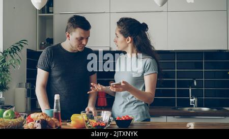 young curly caucasian woman showing something unpleasant in husband's phone while he is cooking , they are shouting and quarrelling, man is angry and irritated Stock Photo