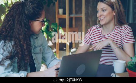 Young female enterpreneurs sitting together at table and talking emotionally. Women are wearing casual clothes and stylish accessories. Loft office in backfround. Stock Photo