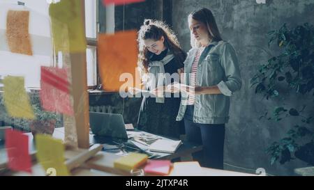 Cheerful designers are standing near table in loft style office and choosing photos for project. Young women are having friendly relations, they are chatting, laughing and sharing ideas. Stock Photo