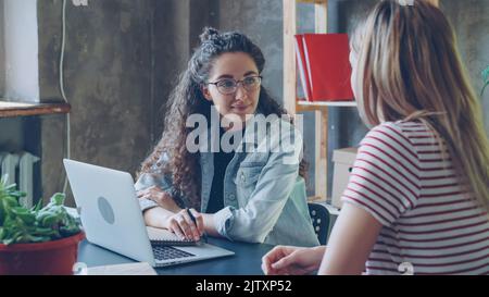 Young female enterpreneur is talking to her attractive partner in modern office while sitting at desk together. Women are planning new project. Successful cooperation concept. Stock Photo