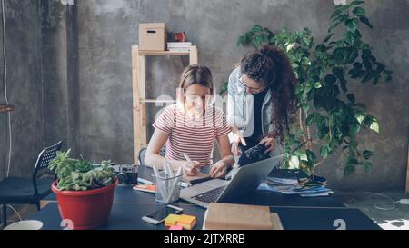 Young designer is drawing images in notebook sitting at table while female photographer is coming to her with camera. Women start discussing new project and watching photos on screen. Stock Photo
