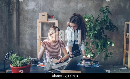 Young creative colleagues are planning design, drawing layout in notebook and watching photos on camera screen. Women are busy working on important project. Desk with office supplies in foreground. Stock Photo