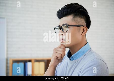 Young pensive businessman in eyeglasses standing at office Stock Photo