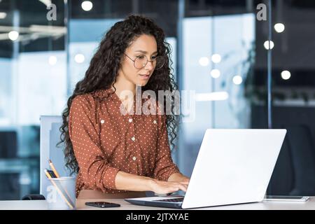 Beautiful indian woman programmer web developer focused and confident working on laptop writing code programming business woman worker in modern office working in casual clothes. Stock Photo