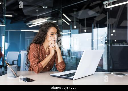 Pensive and focused female programmer web developer working in modern product development company Hispanic woman sitting and thinking using laptop to write code. Stock Photo