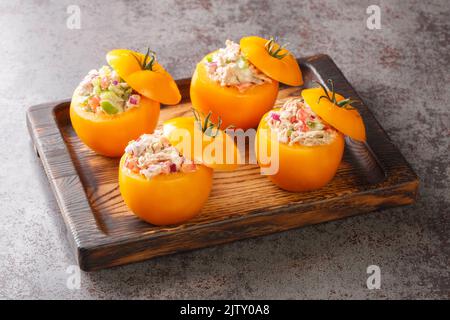 Ripe tomatoes stuffed with a seasonal salad of canned tuna, bell peppers, onions and greens close-up on a wooden tray on the table. Horizontal Stock Photo