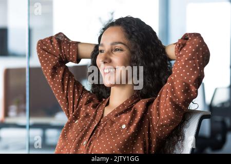 Close-up photo of a young beautiful business woman entrepreneur resting in the office looking out the window with her hands behind her head, businesswoman working on a laptop smiling and dreaming. Stock Photo