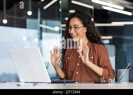 Online meeting video call young beautiful hispanic woman working in modern office, businesswoman using laptop to communicate with colleagues, female worker smiling and saying hello to web camera. Stock Photo