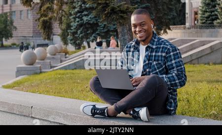 Successful busy african smiling man afro guy student male freelancer programmer designer in wireless headphones sitting on terrace in city in park Stock Photo