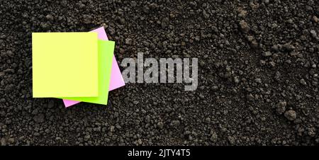 Yellow sticky note blank paper flat soil organic earth texture. Colors note paper square on black soil texture ground close up. Mockup on ground soil Stock Photo