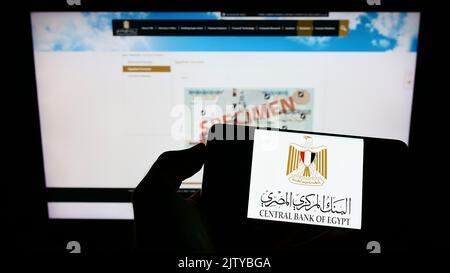 Person holding cellphone with logo of monetary authority Central Bank of Egypt (CBE) on screen in front of webpage. Focus on phone display. Stock Photo