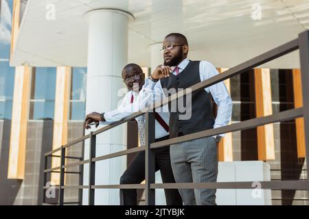 Portrait of two dark-skinned businessmen talking in the background of a modern building exterior. Friendly meeting outdoors Stock Photo