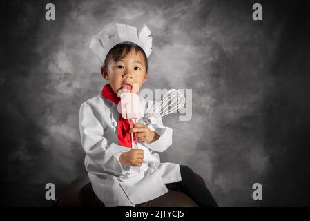 A toddler girl in chef cook attire portrait is licking a plastic spatula while holding a whisk. Stock Photo