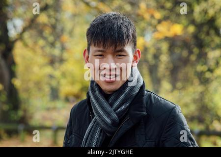 A portrait of a young, Chinese man standing outdoors in nature in Northumberland, North East England during Autumn. He is smiling while looking at the Stock Photo
