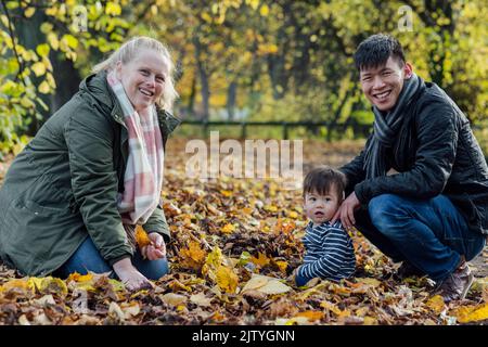 A family crouching down and playing with fallen leaves in nature in Northumberland, North East England. They are all looking at the camera and smiling Stock Photo