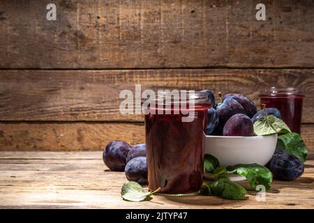 Red blue plum jam in small jar. Homemade autumn plum jam with fresh fruits. Fall preparations and canning on wooden table background copy space Stock Photo