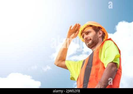A portrait of a worker stands with confidence in an orange work suit and safety helmet against the sky. Smart industrial worker work concept. Stock Photo
