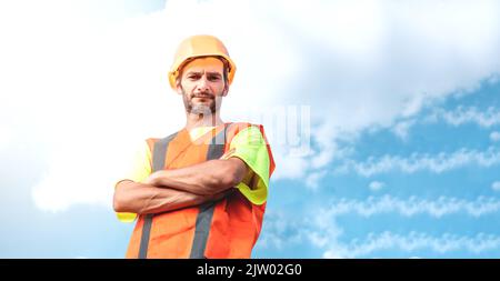 A portrait of a worker stands with confidence in an orange work suit and safety helmet against the sky. Smart industrial worker work concept. Stock Photo