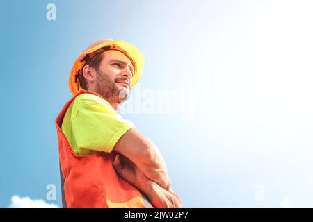 A portrait of a worker stands with confidence in an orange work suit and safety helmet against the sky. Smart industrial worker work concept. Stock Photo