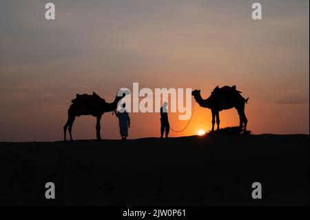 Silhouette of two cameleers and their camels at sand dunes of Thar desert, Rajasthan, India. Cloud with setting sun, sky in the background. Stock Photo