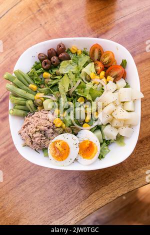 Tuna salad, egg, potato, lettuce, onion, tomato, corn, black olives and asparagus. Served in a white bowl on a wooden table. Healthy meal. Stock Photo