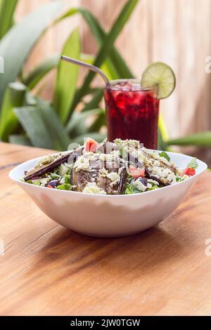 Eggplant salad, parmesan cheese, olives and lettuce served in a white bowl on a wooden table in a restaurant. Accompanied by a glass of red juice. Hea Stock Photo