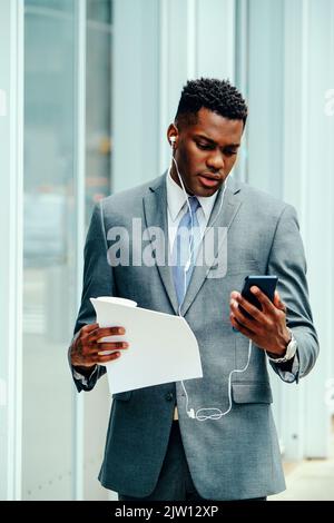 Young adult businessman using smartphone outside wearing suit, tie and headphones holding documents Stock Photo