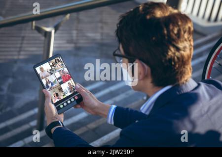 Multiracial business people on video call with caucasian businessman in mask sitting on steps Stock Photo