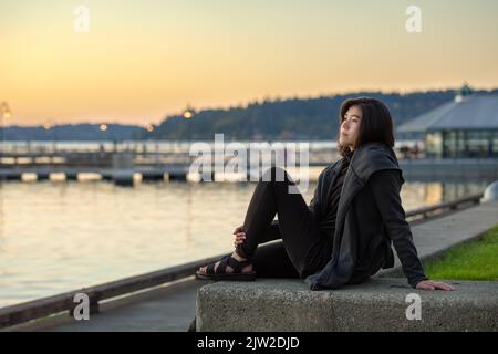 Smiling teen girl or young adult female in gray jacket sitting outdoors by lake enjoying colorful sunset on cool evening Stock Photo