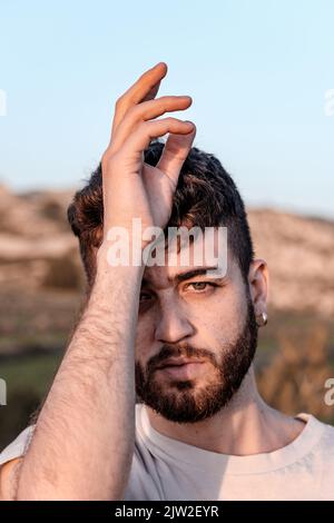 Calm bearded male in white t shirt touching Crown chakra and looking at camera during yoga session against hill and blue sky Stock Photo