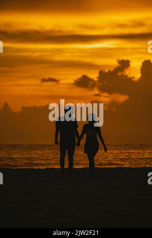 Back view silhouette of anonymous loving couple holding hands and walking on beach towards wavy sea against scenic sunset sky in Clearwater Stock Photo