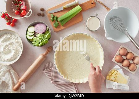 Top view of anonymous female using fork to dock holes in raw quiche crust near kitchenware and fresh ingredients on table in kitchen Stock Photo