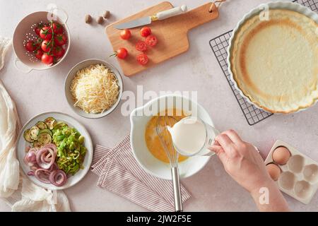 Top view of anonymous female pouring fresh milk into bowl with eggs near various ingredients while preparing quiche on table at home Stock Photo