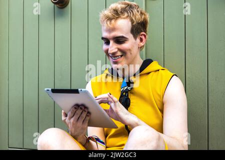 Young smiling transgender man in yellow jumpsuit browsing tablet while sitting near house in evening on city street Stock Photo