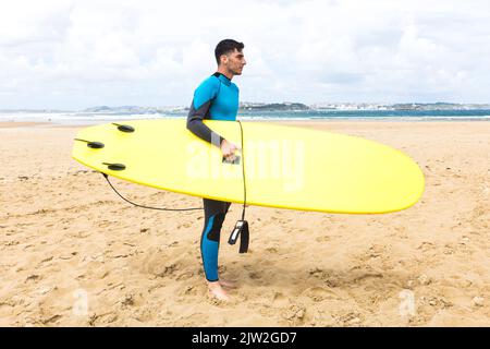 Side view full body of thoughtful young Hispanic male surfer with dark hair and in wetsuit smiling while standing with yellow surfboard on sandy beach Stock Photo