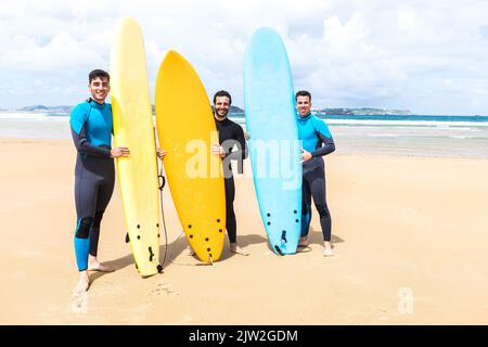 Full body of positive young Hispanic male best friends in wetsuits smiling and looking at camera while standing near blue and yellow surfboards on san Stock Photo