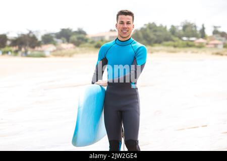 Full body of cheerful young Hispanic male surfer with dark hair and in wetsuit smiling while standing with blue surfboard on sandy beach Stock Photo