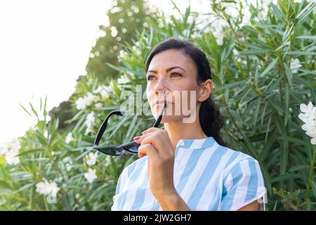 Middle aged female with long dark hair in casual clothes looking away while standing in park with sunglasses in hand Stock Photo