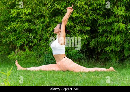 Full body side view of sportive female practicing Hanumanasana posture with raised arms on lawn during yoga training in park Stock Photo