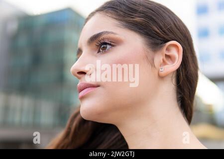 Side view of gorgeous young woman with long brown hair in stylish outfit smiling looking away in city Stock Photo